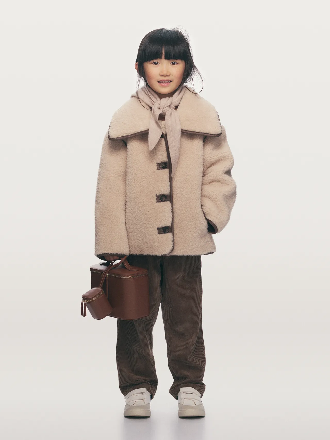 Child wearing a beige shearling coat holding a leather bucket bag.